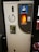 A bottle recycling machine with a slot for inserting bottles, a circular sign indicating recycling, and a lit display. An information sheet with text in a foreign language is attached to the front.