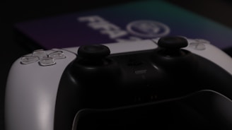 A close-up of a video game controller with a sleek design featuring white and black colors. The focus is on the analog sticks and buttons, with partial visibility of a console or game in the background displaying blurred text.