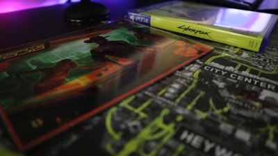 A video game case and associated artwork are displayed on a table. The game case is labeled for a PlayStation 4, and the artwork features a futuristic theme with bold neon colors. The background includes a city map with highlighted text.