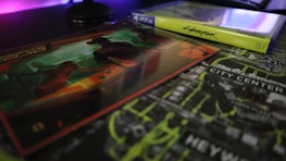 A video game case and associated artwork are displayed on a table. The game case is labeled for a PlayStation 4, and the artwork features a futuristic theme with bold neon colors. The background includes a city map with highlighted text.