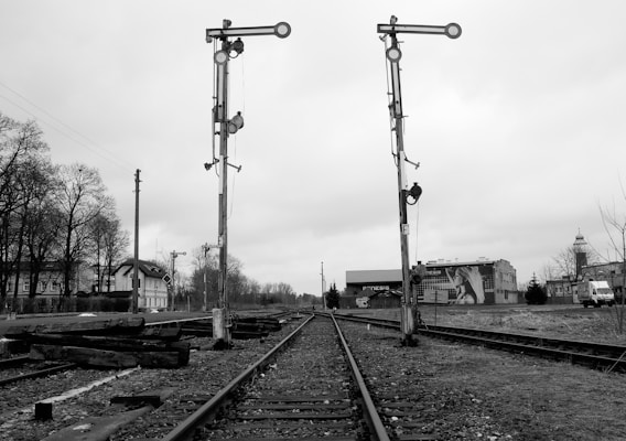 A black and white photograph depicting two old-style railway signal posts on the side of train tracks. The tracks extend into the distance, surrounded by bare trees and buildings. The sky is overcast, adding to a somber mood. An old station building and a vehicle can be seen in the background.