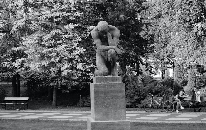 A stone sculpture stands prominently on a pedestal in a park setting, surrounded by lush trees and greenery. The sculpture depicts a human figure in a contemplative pose. Nearby, two people sit on a bench next to a bicycle, with a paved path running through the park.