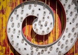 A close-up of a vintage marquee sign featuring circular patterns. The sign is adorned with numerous small, round light bulbs arranged in a spiral, many of which appear to be aged or missing. The backdrop consists of a red and yellow, weathered surface with a scratched and rusted appearance.