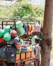 A street vendor selling a variety of colorful plastic household items from the back of a small truck. Various kitchenware and containers, including baskets, buckets, and strainers, are stacked and hung for display. A young boy stands nearby, observing the items.