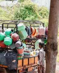 A street vendor selling a variety of colorful plastic household items from the back of a small truck. Various kitchenware and containers, including baskets, buckets, and strainers, are stacked and hung for display. A young boy stands nearby, observing the items.