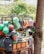 A street vendor selling a variety of colorful plastic household items from the back of a small truck. Various kitchenware and containers, including baskets, buckets, and strainers, are stacked and hung for display. A young boy stands nearby, observing the items.
