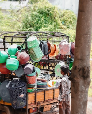 A street vendor selling a variety of colorful plastic household items from the back of a small truck. Various kitchenware and containers, including baskets, buckets, and strainers, are stacked and hung for display. A young boy stands nearby, observing the items.