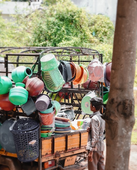 A street vendor selling a variety of colorful plastic household items from the back of a small truck. Various kitchenware and containers, including baskets, buckets, and strainers, are stacked and hung for display. A young boy stands nearby, observing the items.