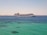 A large cruise ship sails across a calm, expansive ocean with a distant coastline visible in the background under a clear sky.