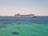 A large cruise ship sails across a calm, expansive ocean with a distant coastline visible in the background under a clear sky.