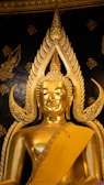 An ornate golden Buddha statue with intricate details set against a dark, decorated background. The statue features elaborate designs around the head and is draped in a golden robe.