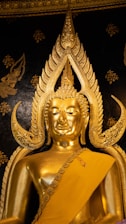 An ornate golden Buddha statue with intricate details set against a dark, decorated background. The statue features elaborate designs around the head and is draped in a golden robe.