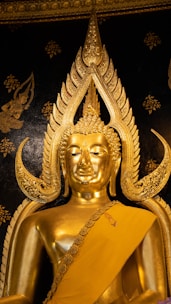 An ornate golden Buddha statue with intricate details set against a dark, decorated background. The statue features elaborate designs around the head and is draped in a golden robe.