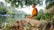 A person in an orange robe sits peacefully on a grassy riverbank, surrounded by lush greenery and rocks. The serene river reflects the trees, creating a calm and tranquil atmosphere. The individual appears to be meditating, embodying a sense of calmness and introspection.