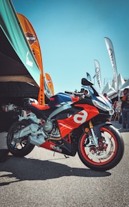 A sporty red and silver motorcycle with the brand name Aprilia prominently displayed is parked on a paved area. Several colorful banners and a large canopy are situated in the background, with people gathered under the tents.