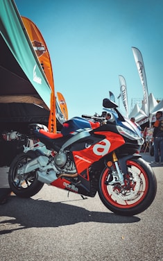 A sporty red and silver motorcycle with the brand name Aprilia prominently displayed is parked on a paved area. Several colorful banners and a large canopy are situated in the background, with people gathered under the tents.
