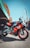 A sporty red and silver motorcycle with the brand name Aprilia prominently displayed is parked on a paved area. Several colorful banners and a large canopy are situated in the background, with people gathered under the tents.