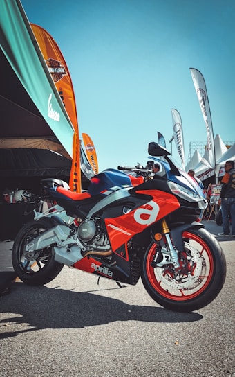 A sporty red and silver motorcycle with the brand name Aprilia prominently displayed is parked on a paved area. Several colorful banners and a large canopy are situated in the background, with people gathered under the tents.