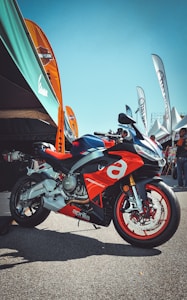 A sporty red and silver motorcycle with the brand name Aprilia prominently displayed is parked on a paved area. Several colorful banners and a large canopy are situated in the background, with people gathered under the tents.