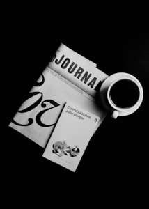 A black and white composition featuring a journal, the cover of John Berger's 'Confabulations', and a cup of coffee. The journal is partially visible, showing the letters 'JOURNAL'. The book cover displays a small illustration. The coffee cup is white with a visible dark coffee inside, placed on the right.