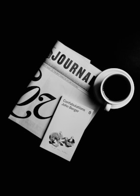 A black and white composition featuring a journal, the cover of John Berger's 'Confabulations', and a cup of coffee. The journal is partially visible, showing the letters 'JOURNAL'. The book cover displays a small illustration. The coffee cup is white with a visible dark coffee inside, placed on the right.