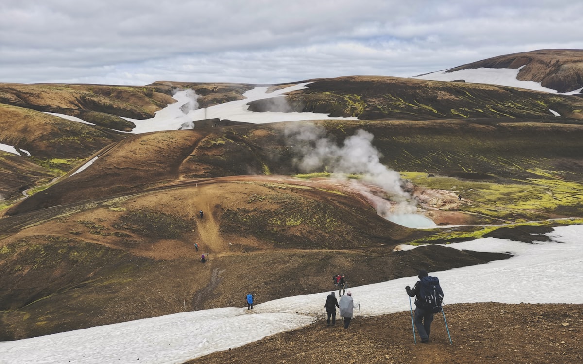 Hikers crossing a snow patch below steaming geothermal vents and multicolored rhyolite hills in the Icelandic highlands