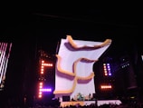A large, illuminated stage with a dynamic, curving light structure dominates the scene, surrounded by colorful lights and electronic displays. The audience is visible in the foreground, creating a lively concert atmosphere.
