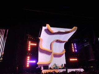 A large, illuminated stage with a dynamic, curving light structure dominates the scene, surrounded by colorful lights and electronic displays. The audience is visible in the foreground, creating a lively concert atmosphere.