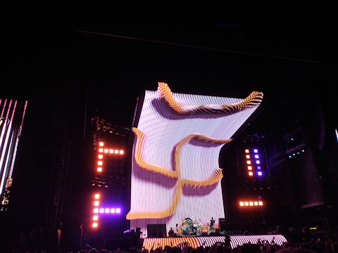 A large, illuminated stage with a dynamic, curving light structure dominates the scene, surrounded by colorful lights and electronic displays. The audience is visible in the foreground, creating a lively concert atmosphere.