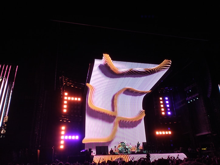 A large, illuminated stage with a dynamic, curving light structure dominates the scene, surrounded by colorful lights and electronic displays. The audience is visible in the foreground, creating a lively concert atmosphere.