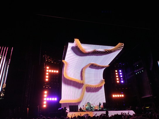 A large, illuminated stage with a dynamic, curving light structure dominates the scene, surrounded by colorful lights and electronic displays. The audience is visible in the foreground, creating a lively concert atmosphere.