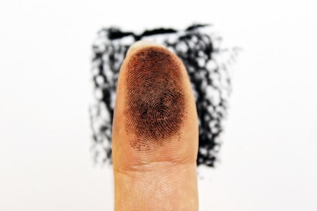 A close-up of a fingertip with a visible fingerprint pattern. The background is blurred and appears to have a dark ink stamp or smudge behind the finger.