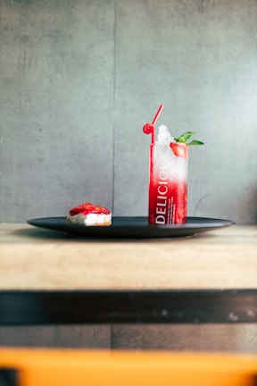 A tall glass filled with a red icy beverage sits on a black tray. The drink has a red straw and is garnished with mint leaves. Next to the drink is a small dessert topped with red strawberries.