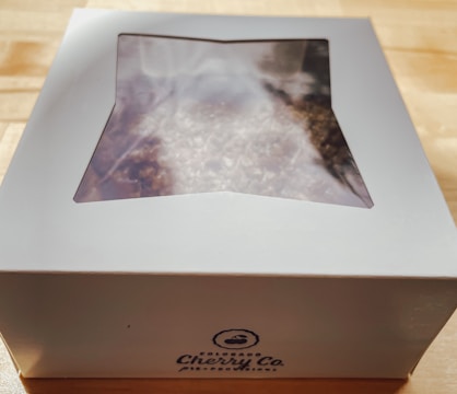A white box labeled 'Colorado Cherry Co.' is on a wooden surface. The box has a clear window on top, revealing a partially visible baked good inside, possibly topped with sugar.