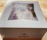 A white box labeled 'Colorado Cherry Co.' is on a wooden surface. The box has a clear window on top, revealing a partially visible baked good inside, possibly topped with sugar.