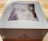 A white box labeled 'Colorado Cherry Co.' is on a wooden surface. The box has a clear window on top, revealing a partially visible baked good inside, possibly topped with sugar.