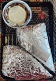 Two items wrapped in silver aluminum foil are placed inside a black plastic tray with geometric patterns. A small plastic cup containing a light-colored sauce with a clear lid is positioned above the foil-wrapped items.