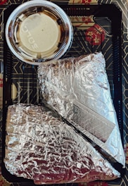 Two items wrapped in silver aluminum foil are placed inside a black plastic tray with geometric patterns. A small plastic cup containing a light-colored sauce with a clear lid is positioned above the foil-wrapped items.