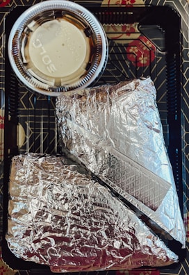 Two items wrapped in silver aluminum foil are placed inside a black plastic tray with geometric patterns. A small plastic cup containing a light-colored sauce with a clear lid is positioned above the foil-wrapped items.