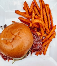 A freshly prepared hamburger placed on a white plate, accompanied by a generous serving of crispy, golden brown fries. The hamburger is topped with a soft, shiny bun and contains what appears to be a thick patty glazed with a glossy reddish sauce.