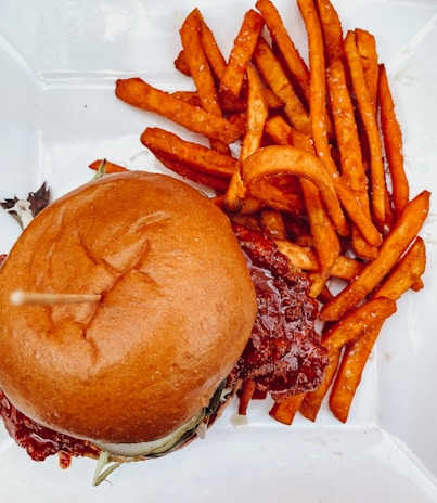 A freshly prepared hamburger placed on a white plate, accompanied by a generous serving of crispy, golden brown fries. The hamburger is topped with a soft, shiny bun and contains what appears to be a thick patty glazed with a glossy reddish sauce.