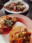 Three plates of tacos, each topped with a variety of ingredients including meats, chopped onions, cilantro, sliced radishes, and pickled jalapeños. The tacos are presented on corn tortillas and are arranged on simple white plates, sitting on a dark surface.