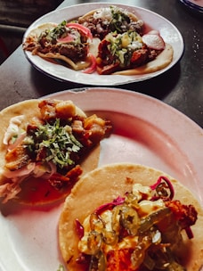 Three plates of tacos, each topped with a variety of ingredients including meats, chopped onions, cilantro, sliced radishes, and pickled jalapeños. The tacos are presented on corn tortillas and are arranged on simple white plates, sitting on a dark surface.