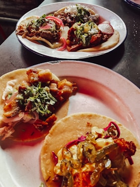 Three plates of tacos, each topped with a variety of ingredients including meats, chopped onions, cilantro, sliced radishes, and pickled jalapeños. The tacos are presented on corn tortillas and are arranged on simple white plates, sitting on a dark surface.