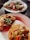 Three plates of tacos, each topped with a variety of ingredients including meats, chopped onions, cilantro, sliced radishes, and pickled jalapeños. The tacos are presented on corn tortillas and are arranged on simple white plates, sitting on a dark surface.