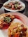 Three plates of tacos, each topped with a variety of ingredients including meats, chopped onions, cilantro, sliced radishes, and pickled jalapeños. The tacos are presented on corn tortillas and are arranged on simple white plates, sitting on a dark surface.