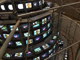 A large installation consisting of multiple rows of vintage television sets stacked in a spiral. These TVs are all displaying various images, some bright and colorful, while others are more subdued. Metal scaffolding surrounds and supports the structure, giving it an industrial appearance. The scene is captured from a higher viewpoint, emphasizing the scale and intricacy of the setup.