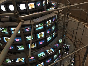 A large installation consisting of multiple rows of vintage television sets stacked in a spiral. These TVs are all displaying various images, some bright and colorful, while others are more subdued. Metal scaffolding surrounds and supports the structure, giving it an industrial appearance. The scene is captured from a higher viewpoint, emphasizing the scale and intricacy of the setup.