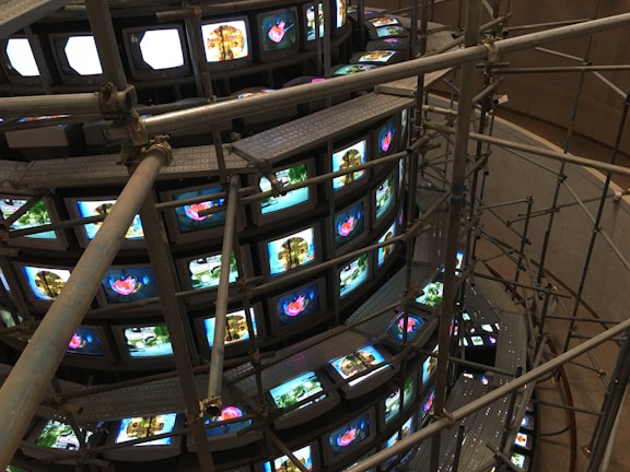 A large installation consisting of multiple rows of vintage television sets stacked in a spiral. These TVs are all displaying various images, some bright and colorful, while others are more subdued. Metal scaffolding surrounds and supports the structure, giving it an industrial appearance. The scene is captured from a higher viewpoint, emphasizing the scale and intricacy of the setup.
