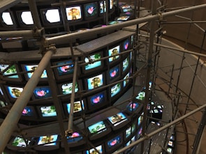 A large installation consisting of multiple rows of vintage television sets stacked in a spiral. These TVs are all displaying various images, some bright and colorful, while others are more subdued. Metal scaffolding surrounds and supports the structure, giving it an industrial appearance. The scene is captured from a higher viewpoint, emphasizing the scale and intricacy of the setup.